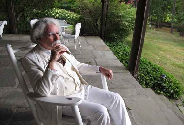 mark dawidziak in a white suit dressed as mark twain sitting in a rocking chair on an outside porch